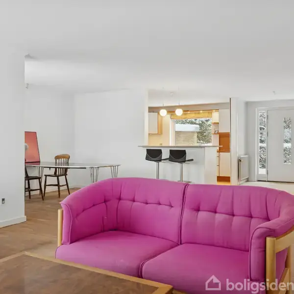 Pink sofa står stille i lys stue med hvidmalede vægge og trægulv. I baggrunden ses spisebordsstole, et åbent køkkenområde og snedækket udsigt gennem glasdøre.