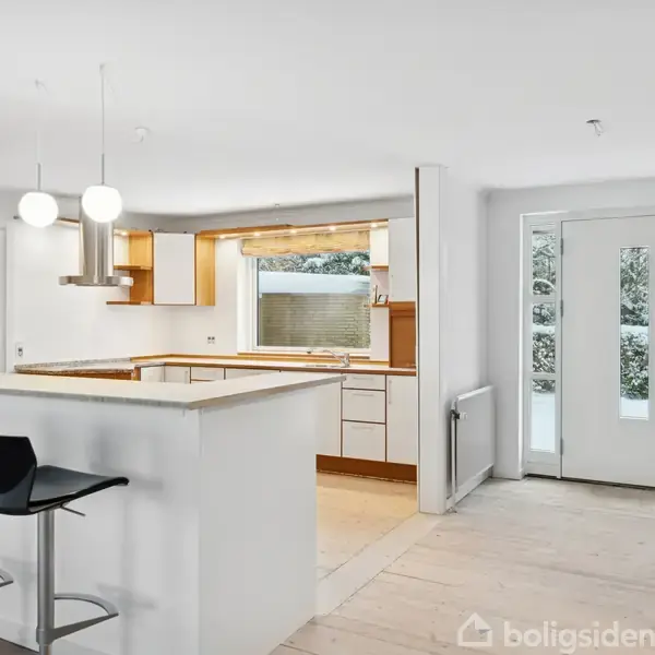 Køkken-ø med to sorte barstole ved siden af. Indgangen til rummet er i baggrunden. Der er store vinduer bag køkkenet, der giver naturligt lys.