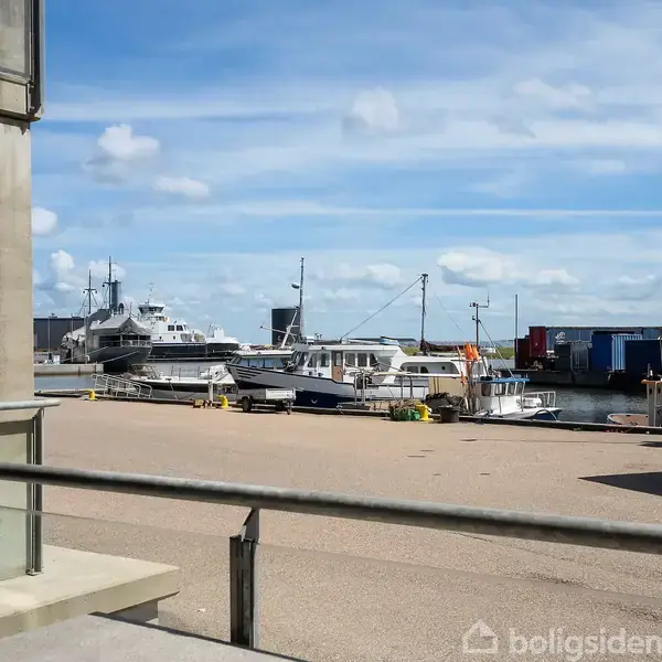 Køkkenalrum med spisebord og seks stole. Moderne køkken til højre med hvidt interiør og blå flisevæg. Sofa med farvede puder bagved. Vindue med udsigt til terrasse i baggrunden.