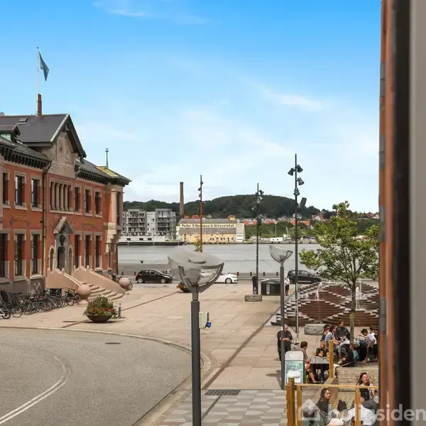 Et rødt murstenshus står stille ved en stille vej, der fører til en havnefront. Folk sidder på bænke tæt på vandet, mens biler er parkeret nær bygningen.