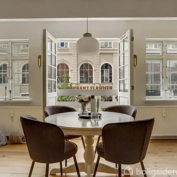 Rundt bord med fire stole står ved åbne dobbeltdøre i en lys spisestue. Udenfor ses en bygning med skiltet "RESTAURANT FLAMMEN". Vinduer omkranser lokalet.