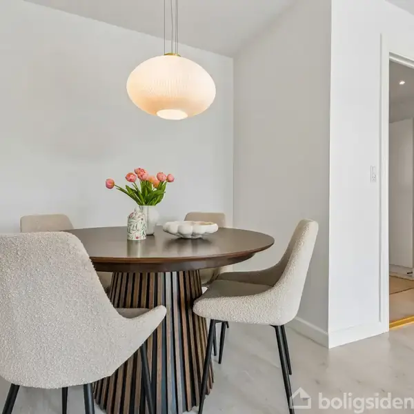 Spisebord med fire polstrede stole står i et lyst rum. En vase med lyserøde tulipaner pynter bordet, og en rund loftslampe hænger over. En glasdør ses i baggrunden.
