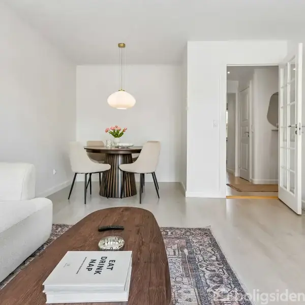 Et moderne stueområde med en hvid sofa, et rektangulært træbord og en bog med teksten "EAT SHOP DRINK MAP." Et rundt spisebord med fire stole i baggrunden. En åben dør fører til en gang.
