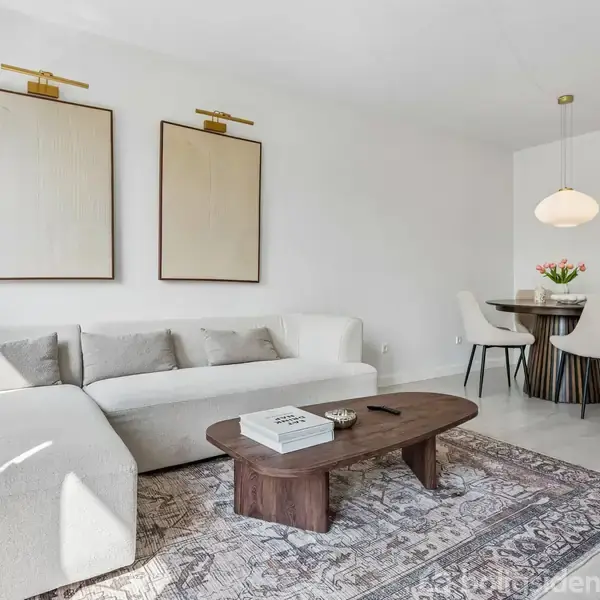 En lys stue med en beige sofa står ved siden af et ovalt, mørkt træbord. På væggen hænger to billeder. Et rundt spisebord med blomster ligger i baggrunden.