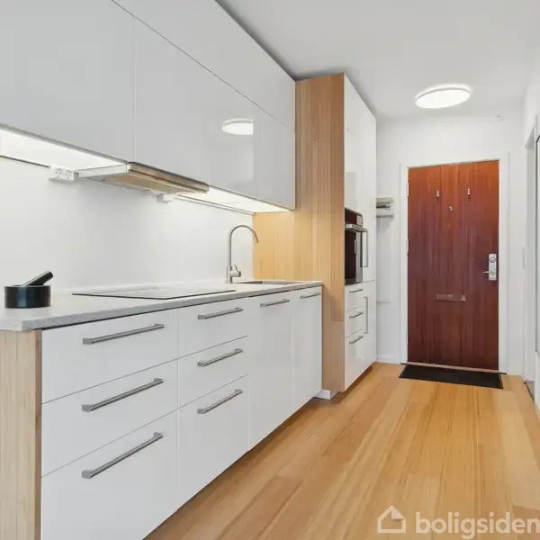 Moderne køkken med hvide skabe og bordplader, hvor en kasserolle står på ovnen. Rummet har træparketgulv og en mørk trædør i baggrunden.