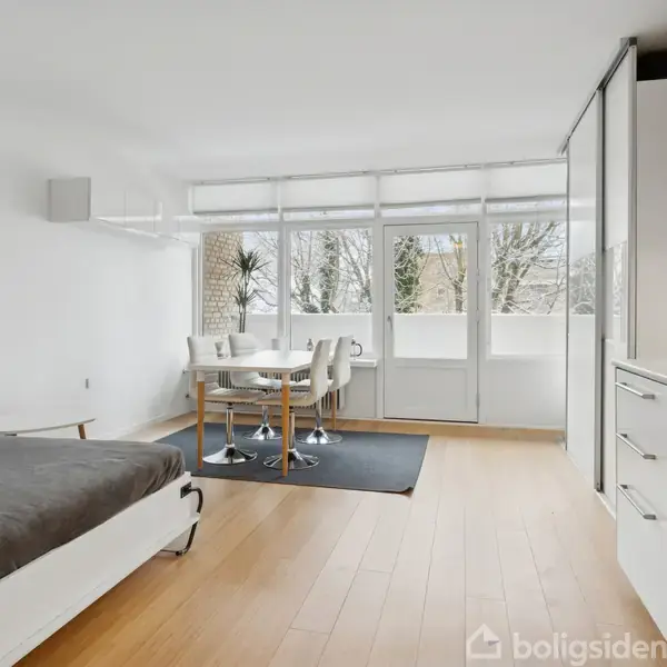En moderne lejlighed med et åbent rum inkluderer en seng og spisebord. Store vinduer i baggrunden giver udsigt til træer, mens et hvidt køkken fylder højre side.