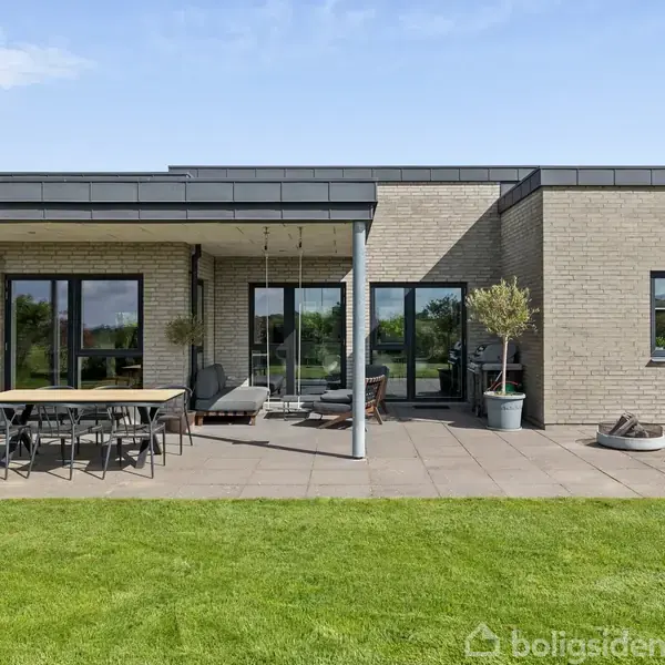 En moderne, enkelt-etage husfacade med store glasdøre, en terrasse med havemøbler og planter, omgivet af en grøn græsplæne under en blå himmel.