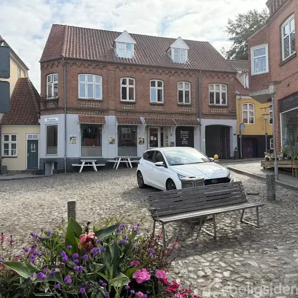 En hvid bil parkerer på en brostensbelagt plads foran en gammel murstensbygning med flere vinduer. Blomster og en bænk ses i forgrunden; omgivet af andre bygninger.