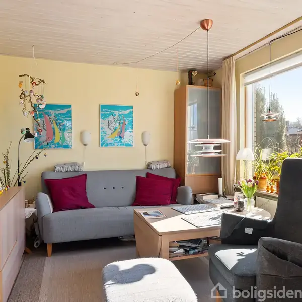 Sofa med røde puder står ved beige væg med kunstplakater. Stue har stort vindue, tv og sofabord. Lysekrone hænger fra loftet. Grønne planter ved vindue.