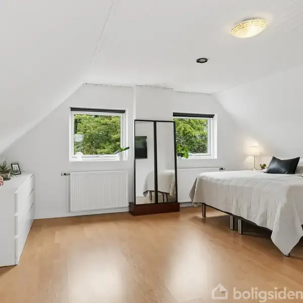 Et soveværelse med en seng og to puder, placeret ved siden af et natbord med en lampe. Der står et stort spejl mellem to vinduer. Lyse trægulve og hvidmalede vægge.