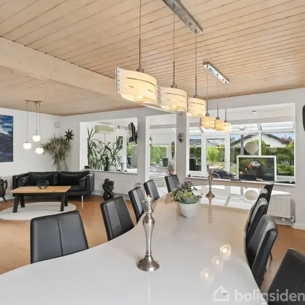 Langt spisebord med sorte stole i moderne stue, dekoreret med planter og kunst på væggen. Store vinduer giver udsigt til have og lyse loftslamper hænger over bordet.