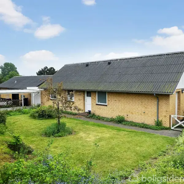 Et gult muret hus med gråt tag står omgivet af en grøn have. Græsplænen foran huset er velplejet med enkelte buske og træer. I baggrunden ses en pergola.