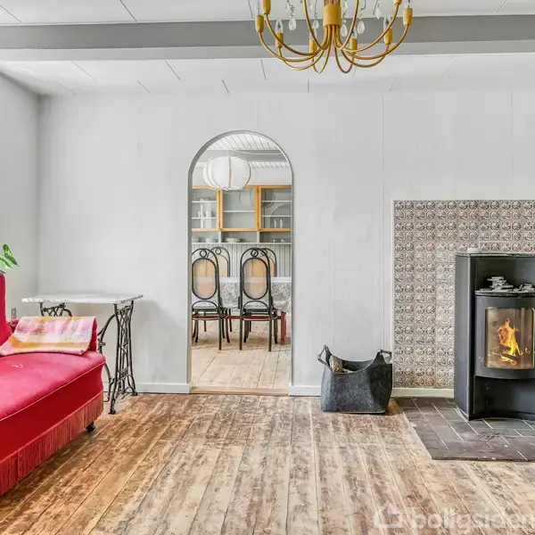 Rød sofa i stue med ild i brændeovn; trægulv og hvid malet væg. Buegang fører til spisestue med stole og skænk. Grøn plante nær vindue.