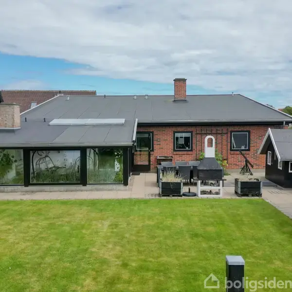 Murstenshus med skrå tag, solpaneler og glaseret udestue; omgivet af terrasse med græsplæne, grill, udemøbler og skur; beliggende i et boligområde.