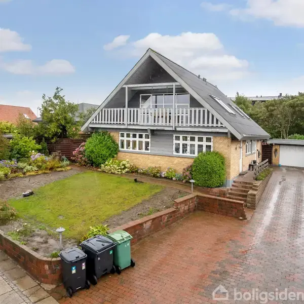 Et hus med en førstesal og en bred balkon ud mod en velplejet have. Flisebelagt indkørsel fører til en garage. Omkringliggende er der grønne træer og andre huse.