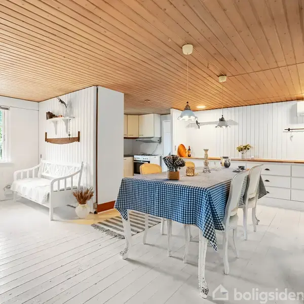 En lys spisestue har et bord med blåternet dug omgivet af hvide stole. Til venstre ses en hvid sofa ved et vindue. Træloft og hvide vægge skaber en hyggelig atmosfære.