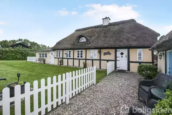 Stråtækt bindingsværkshus med gule vægge og sorte bjælker, omgivet af en hvid stakithegn. En grussti leder til hoveddøren, og en græsplæne er til venstre.