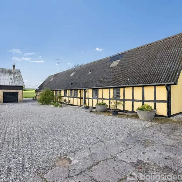 En lang, gul bindingsværksbygning med sort tag står stille på en gårdsplads med småsten. Til venstre ses en mindre hvid bygning. Omgivet af marker og græsplæner.