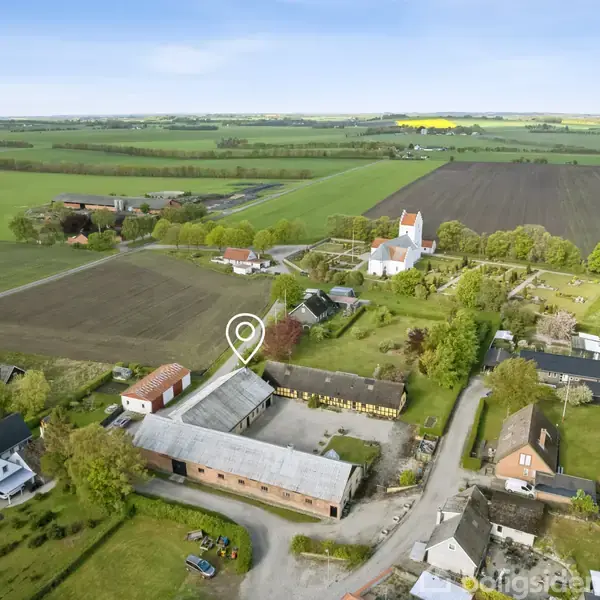 En gård med flere bygninger ligger i et åbent, frodigt landskab. Der er marker og en kirke i nærheden. En cirkelmarkering peger på en af bygningerne.