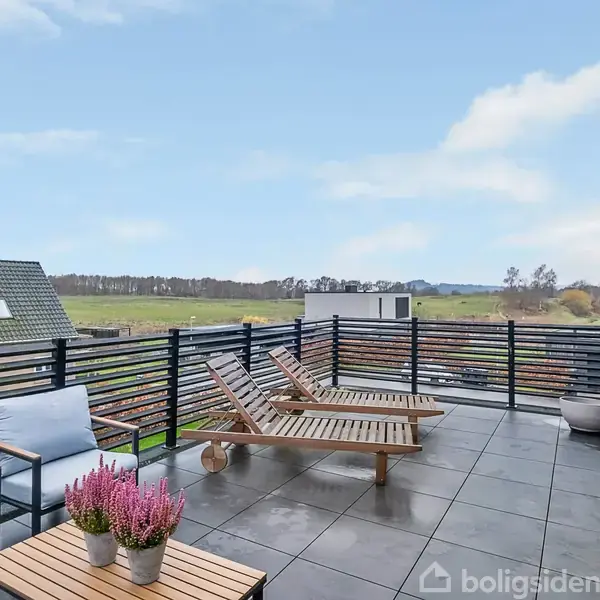 Terrasse med to træliggestole og bord med lilla blomster. Udsigt over grønt landskab og huse. Himlen er blå med nogle skyer.
