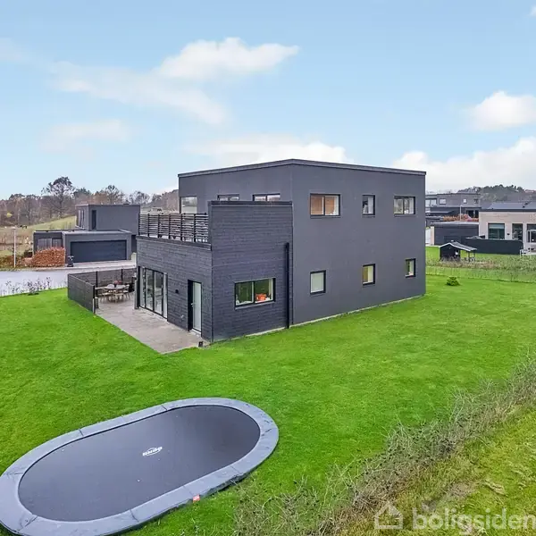 En moderne, sort, kubistisk villa står på en velplejet græsplæne med en nedgravet trampolin foran. Villaen har store vinduer og ligger i et roligt beboelsesområde.