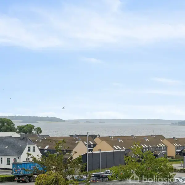 En række huse står stille foran en sø med blå himmel og skyer. En Lidl-butik ses i forgrunden ved vejen.
