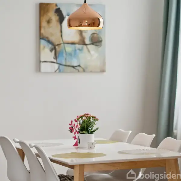 Et spisebord med fire hvide stole står i et lyst rum. En kobberlampe hænger over bordet, og en blomst i en krukke pynter midten. Et abstrakt maleri hænger på væggen.