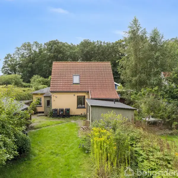 Hus med rødt tegltag står blandt grønt løv, to skraldespande langs væggen. En stor græsplæne strækker sig foran huset, omgivet af træer og buske.
