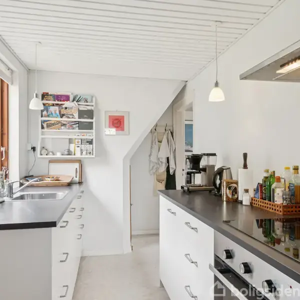 Moderne køkken med hvide skabe, sort bordplade og diverse køkkenmaskiner. En vask er placeret under vinduet. Væghylde med nogle bøger og småting. Lyst interiør med hængelamper.