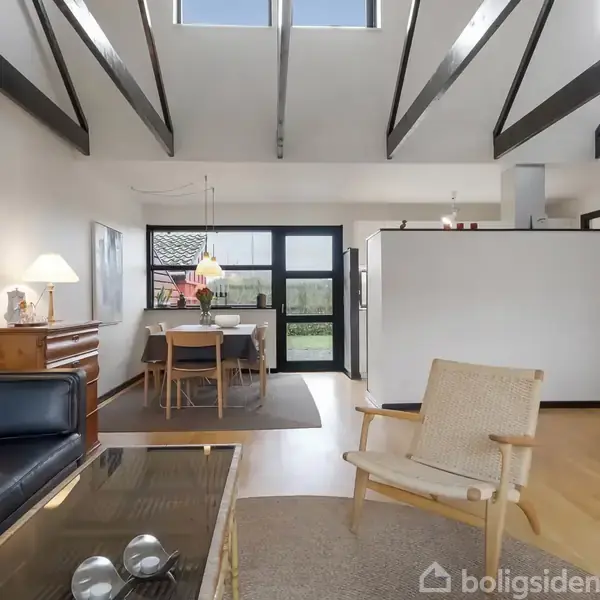 Stue med sofa og lænestol i moderne stil. Spisebord med stole set nær vindue. Åben køkken i baggrunden. Højt til loftet med synlige bjælker og naturligt lysindfald.