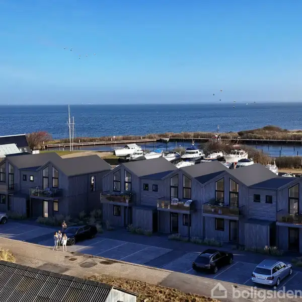 Moderne rækkehuse står stille i en kystnær landsby, omgivet af en parkeringsplads med biler, med udsigt over en lille havn og et bredt, blåt hav i baggrunden.