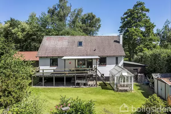 Et hvidt hus med en stor veranda står i en frodig have. Et glasdrivhus ses ved siden af, omgivet af træer og tætte buske.