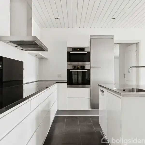 Hvidt køkken med moderne apparater; ovn, mikroovn og køleskab indbygget i skabsmodul. Sort bordplade. Frihængende emhætte over kogeplade. Lys træbeklædning i loft. Vandhane på køkkenø med vask.