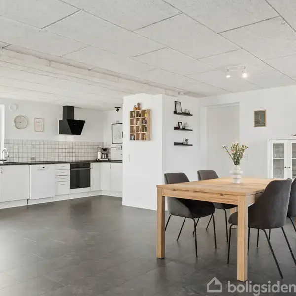 Sort badeværelsesmøbel med håndvask står mod en hvid væg. Over det er et stort spejl. Til venstre ses en bruseniche med mørke fliser. Rummet er lyst og moderne.