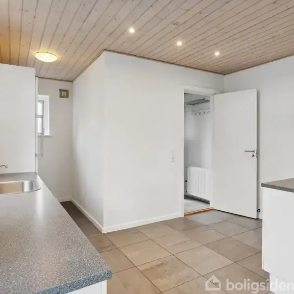 Køkken med grå bordplader og vask, omgivet af hvide skabe. Åben dør fører til en anden del af huset. Loftet er træbeklædt med indbyggede spotlys.