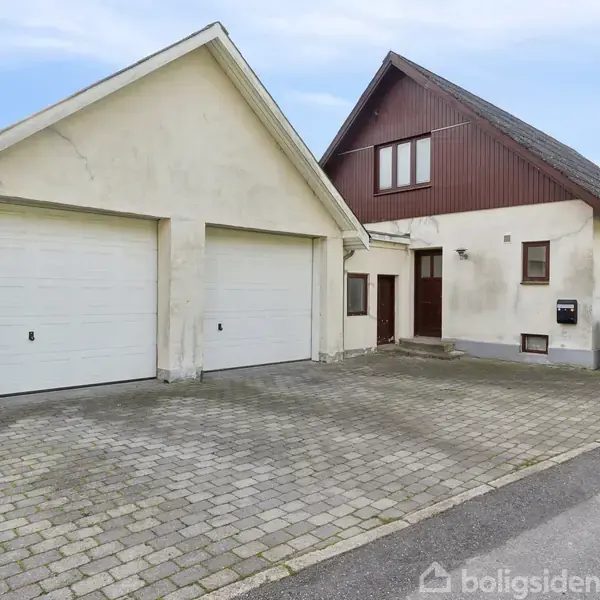 Et hvidt hus med et rødt tag ligger ved siden af en garage med to porte. Det befinder sig i en brostensbelagt indkørsel med grønne træer i baggrunden.