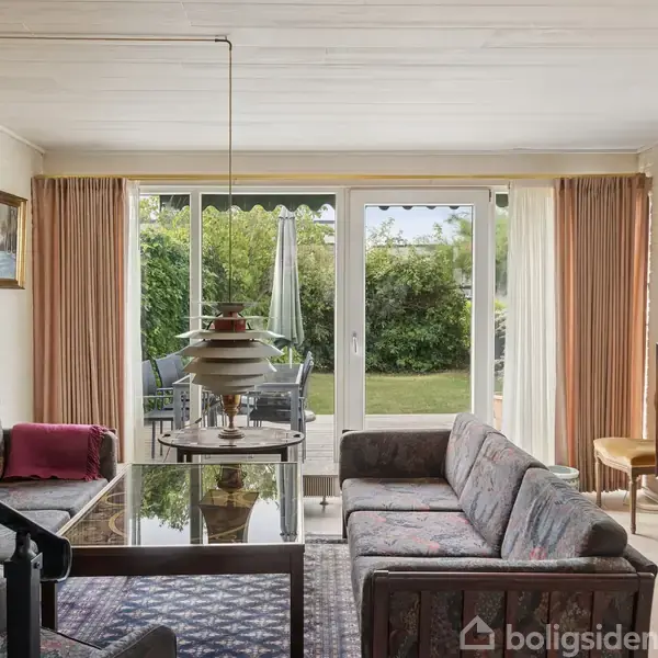 Stue med sofagruppe og sofabord. Store vinduer med udsigt til en have, dekorative malerier på væggene. Der er et rundt ophængt loftlys og en ældre standerlampe.