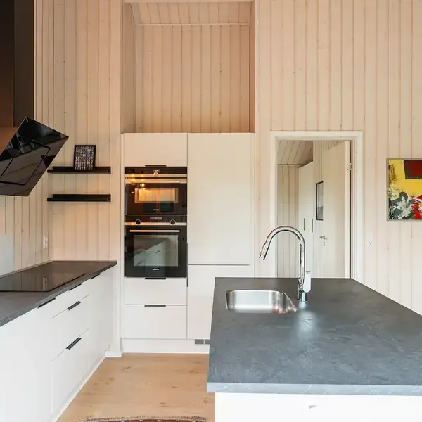 Et moderne køkken med en stor, mørk bordplade, indbyggede hvide skabe og ovne. En dekorativ lampe hænger over bordpladen. Trævægge og en dør fører til et tilstødende rum.