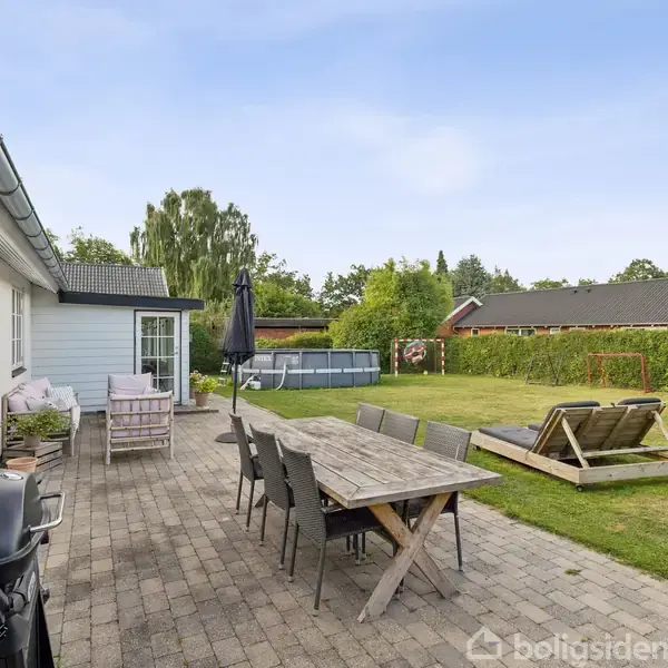 Terrassebord med stole står på en flisebelagt terrasse foran et hus. En grill ses til venstre, mens en græsplæne med gyngeseng er i baggrunden omgivet af grønne planter.
