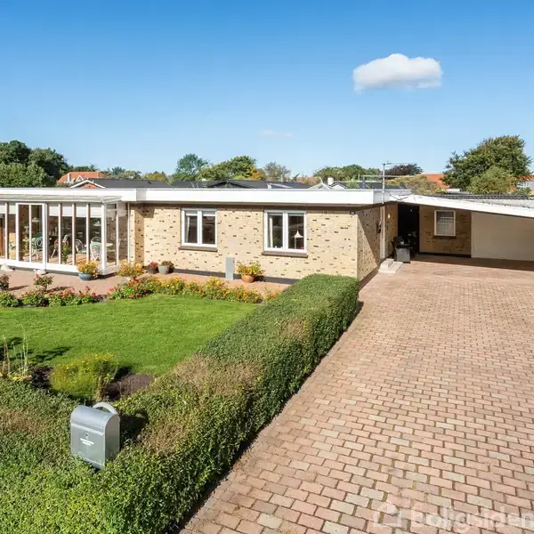 Et en-etagers murstenshus står stille ved siden af en velplejet have og en lang brolagt indkørsel under en blå himmel med en enkelt sky.