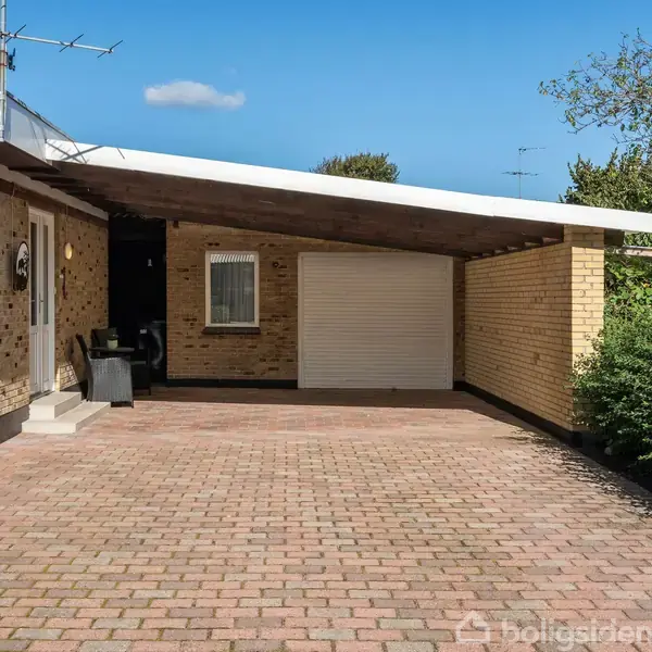 Et murstenshus med fladt tag står i et solrigt miljø. En garageport og en indkørsel med rødbrune fliser leder op til en overbygget indgang. Grønt løv omkranser området.