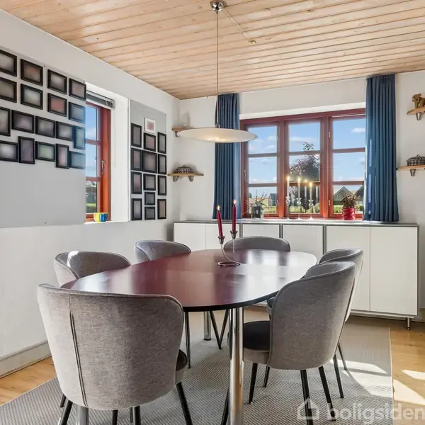 Rundt spisebord med seks polstrede stole står i et lyst rum. Store vinduer med blå gardiner giver udsigt til haven. Væggen er dekoreret med billedrammer.