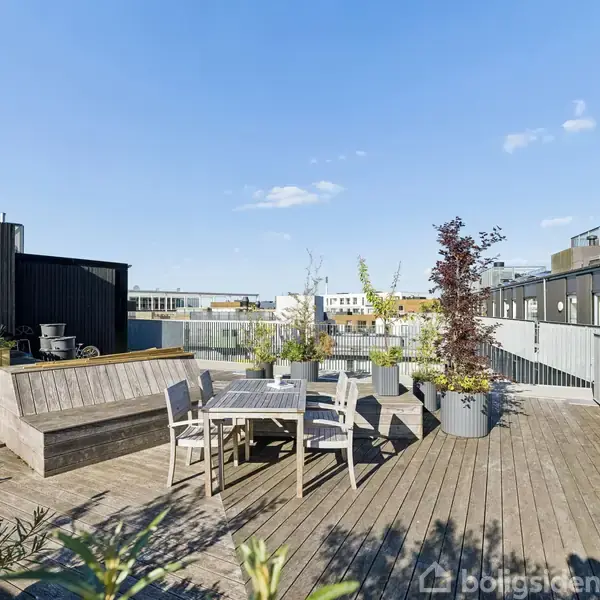 Tagterrasse med et bord og flere stole står på et trægulv. Omkring er der plantekasser med grønt løv under en klar blå himmel i et urbant miljø.