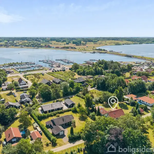 Hus markeret med en cirkel i et villakvarter, omgivet af grønne træer. Nær ved en stor marina ved en flod eller sø, med landskab og marker i baggrunden.