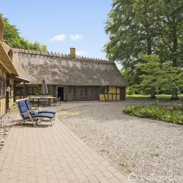 Gule, stråtækte bindingsværkshuse står i en gårdhave med fliser og to liggestole. Omgivet af grønne træer og buske, skaber de en idyllisk, landlig atmosfære.