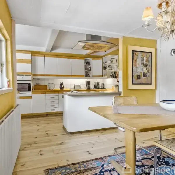 Et spisebord med stole står i et lyst køkken med gule vægge og træelementer. Ovenover hænger en lysekrone. En tæppe dekorerer det lyse trægulv.