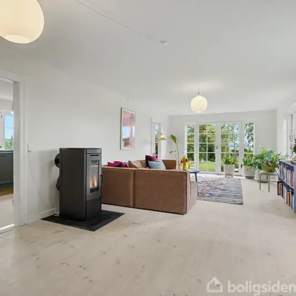 Stue med brændeovn ved siden af en brun sofa. Store vinduer i baggrunden slipper lys ind, og der er planter og en lampe. Køkken er synligt gennem døråbningen.