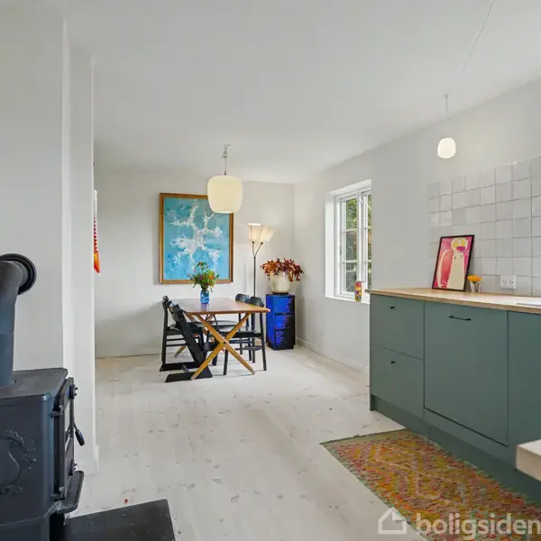 En køkken med en grøn køkkenø og træoverflade, pejs til venstre. Et spisebord med stole under en lampe ses i baggrunden. Vinduer giver udsigt til naturlig omgivelser.