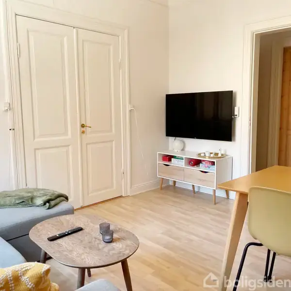 Tv hænger på væggen over et lavt bord med små opbevaringsrum. En sofa og sofabord med fjernbetjening og et stearinlys står ved siden af i et lyst rum. Der er ingen tekst i billedet.