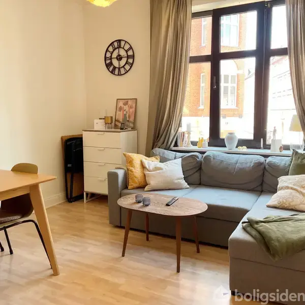En stue indeholder en grå sofa med puder, et sofabord, og et spisebord med stole. Vægur hænger over en kommode ved vinduet med gardiner og udsigt til bygninger.